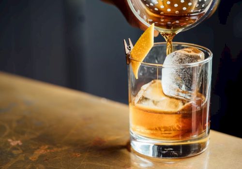 A glass of whiskey on the rocks with a large ice cube and an orange peel garnish being poured into it.