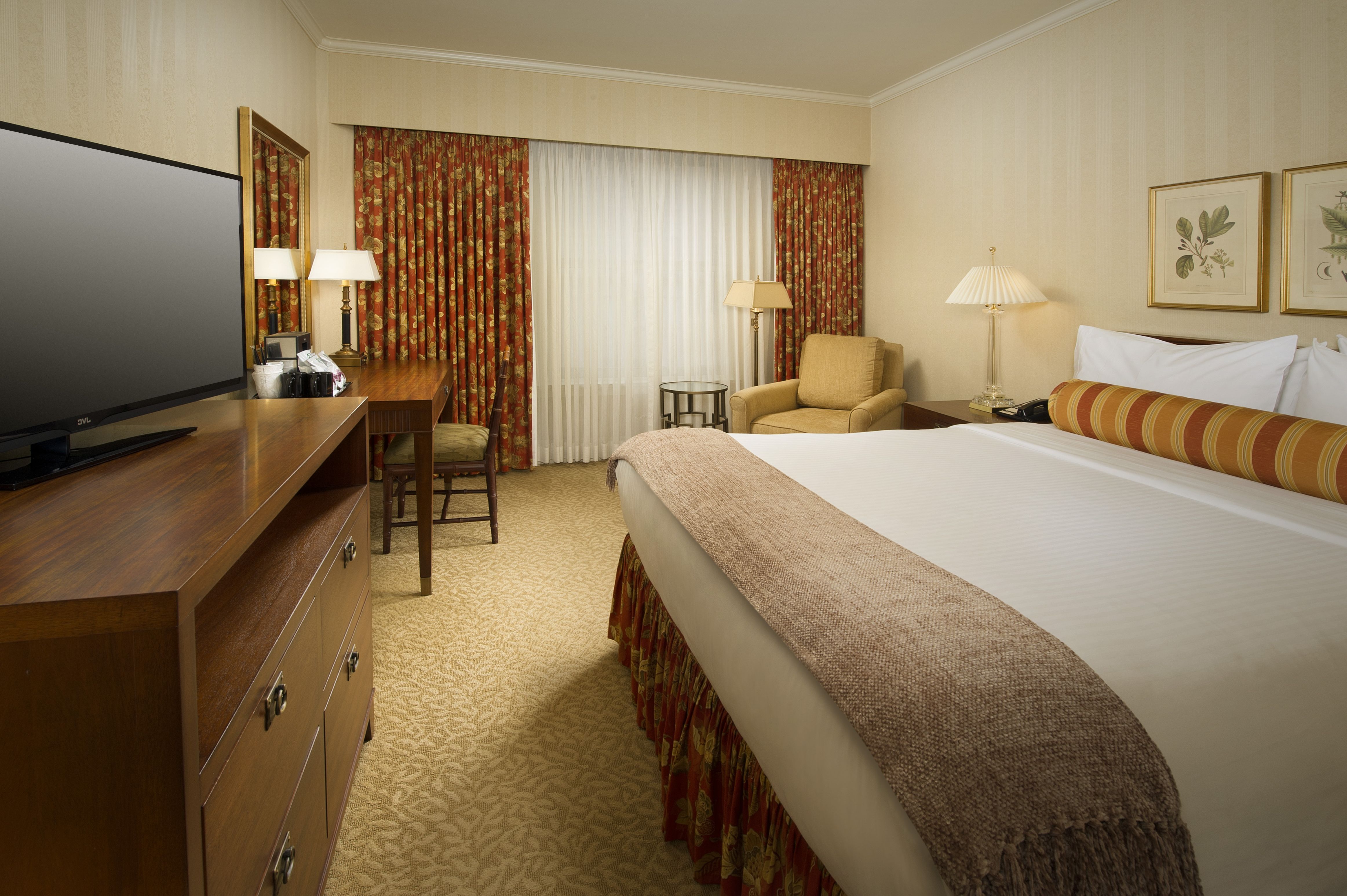 A hotel room with a large bed, a TV on a dresser, a desk and chair, and a window with curtains.
