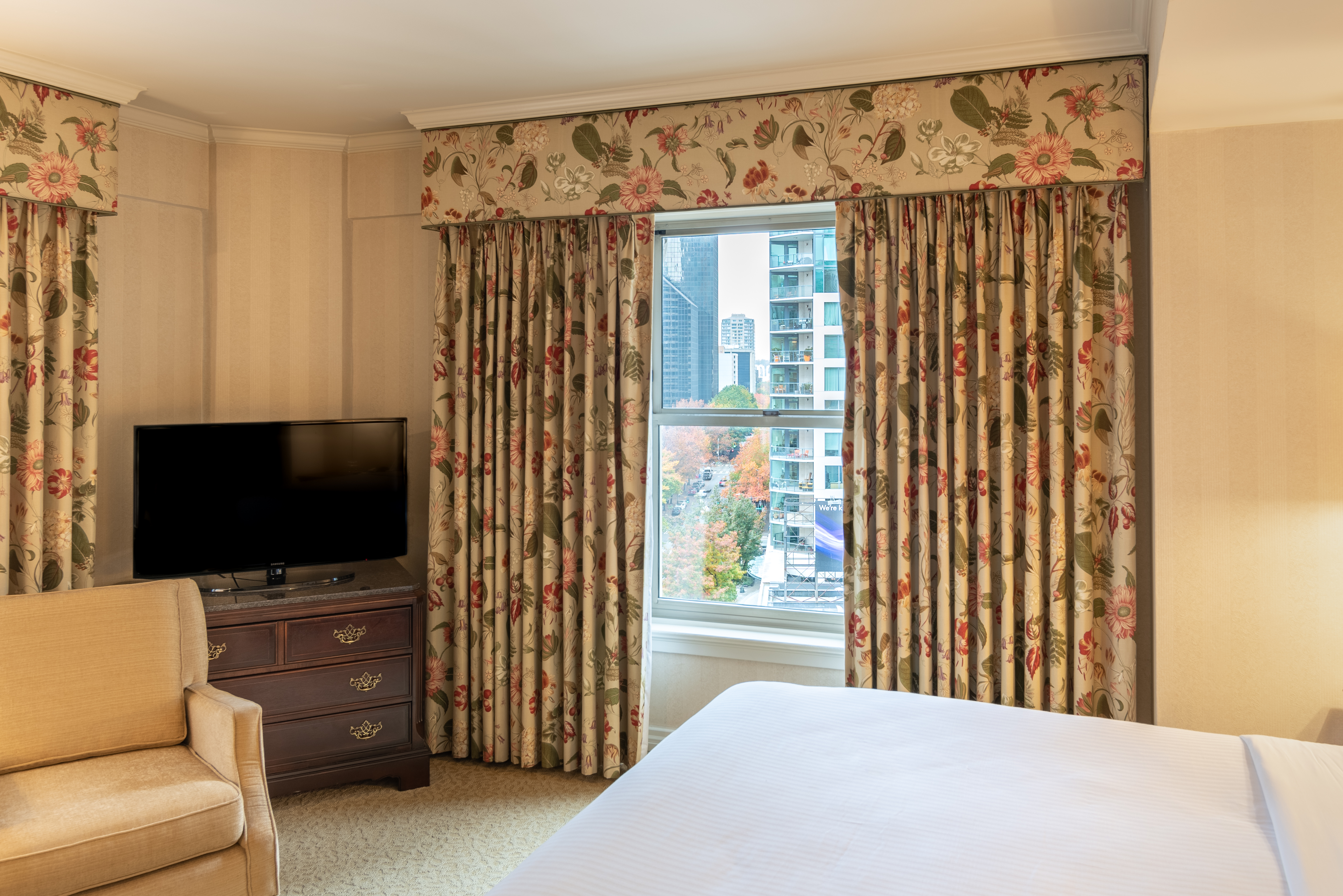 A hotel room with a bed, chair, TV, and window with floral curtains.